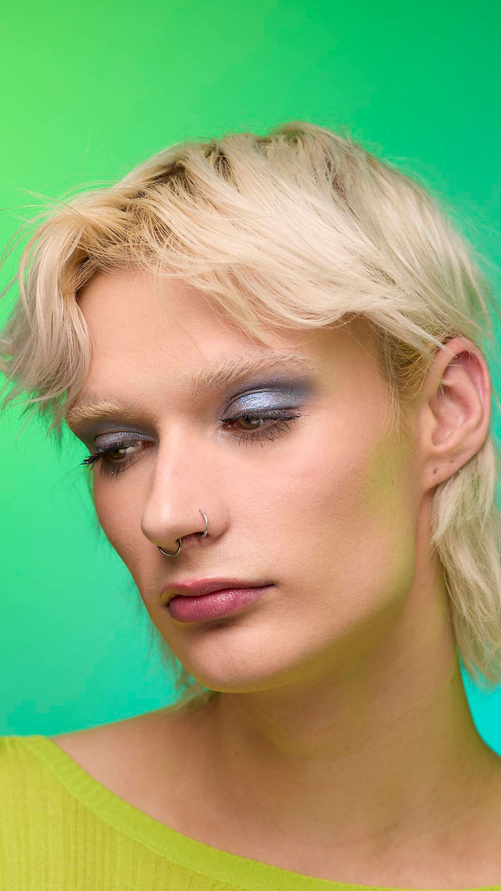 The image shows a close-up of the model's face on a pale blue-to-green gradient background. The model's make-up is done with the focal point being a subtle, midnight-blue, shimmering eyeshadow made by the Nebula Glitter Crumble.
