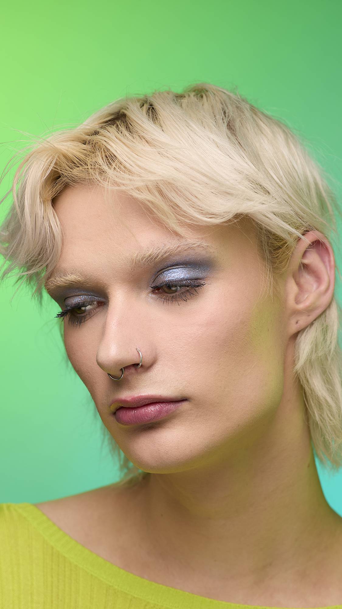 The image shows a close-up of the model's face on a pale blue-to-green gradient background. The model's make-up is done with the focal point being a subtle, midnight-blue, shimmering eyeshadow made by the Nebula Glitter Crumble.