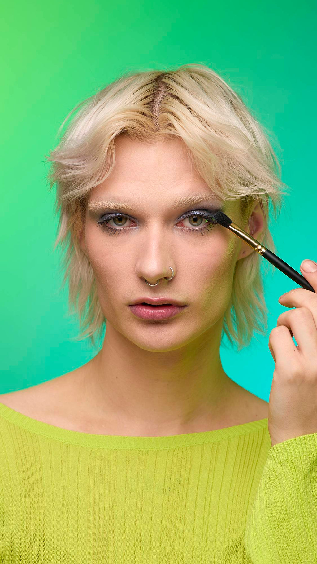 The image shows a close-up of the model's face on a pale blue-to-green gradient background. They are holding a thin make-up brush to apply a subtle, midnight-blue, shimmering eyeshadow of the Nebula Glitter Crumble.