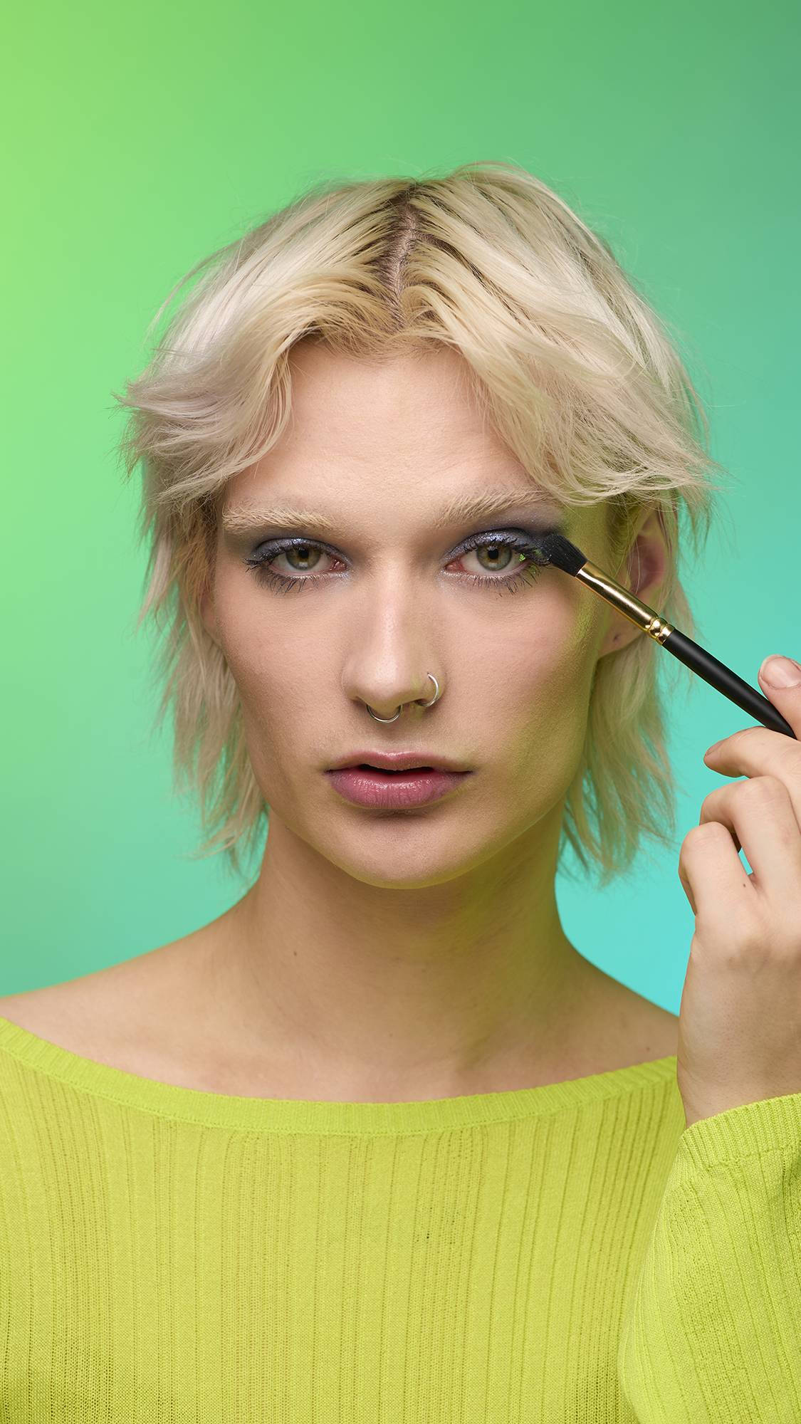 The image shows a close-up of the model's face on a pale blue-to-green gradient background. They are holding a thin make-up brush to apply a subtle, midnight-blue, shimmering eyeshadow of the Nebula Glitter Crumble.