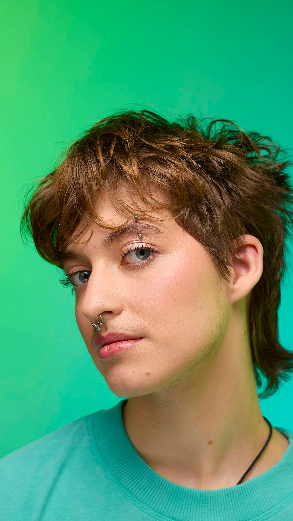 The image shows a close-up of the model's face on a pale blue-to-green gradient background. The model's make-up is done with the focal point being a subtle, iridescent, shimmering eyeshadow made by the New Moon Glitter Crumble.