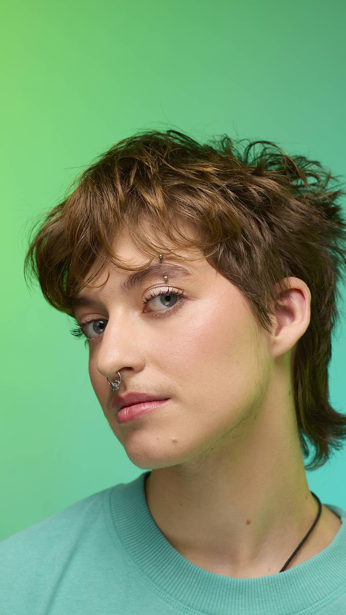 The image shows a close-up of the model's face on a pale blue-to-green gradient background. The model's make-up is done with the focal point being a subtle, iridescent, shimmering eyeshadow made by the New Moon Glitter Crumble.