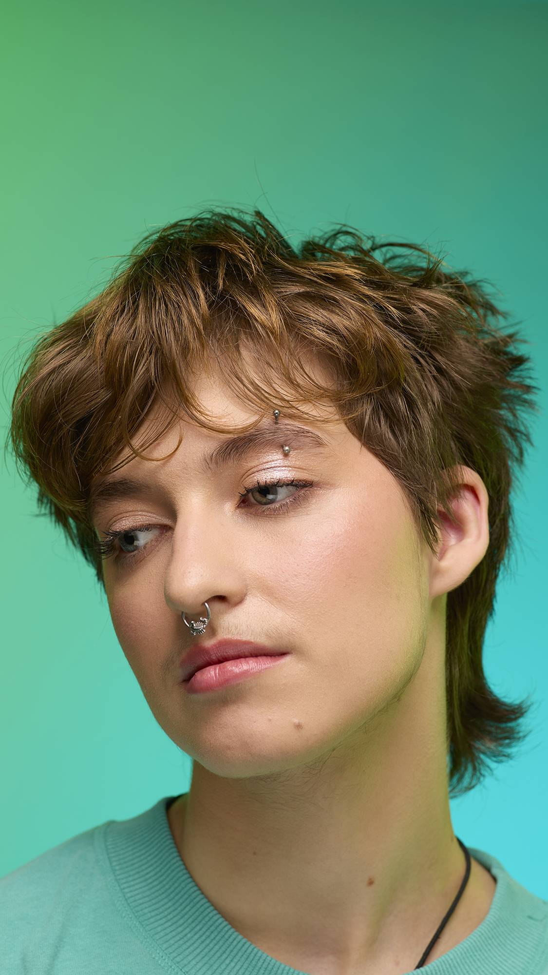 The image shows a close-up of the model's face on a pale blue-to-green gradient background. The model's make-up is done with the focal point being a subtle, iridescent, shimmering eyeshadow made by the New Moon Glitter Crumble.