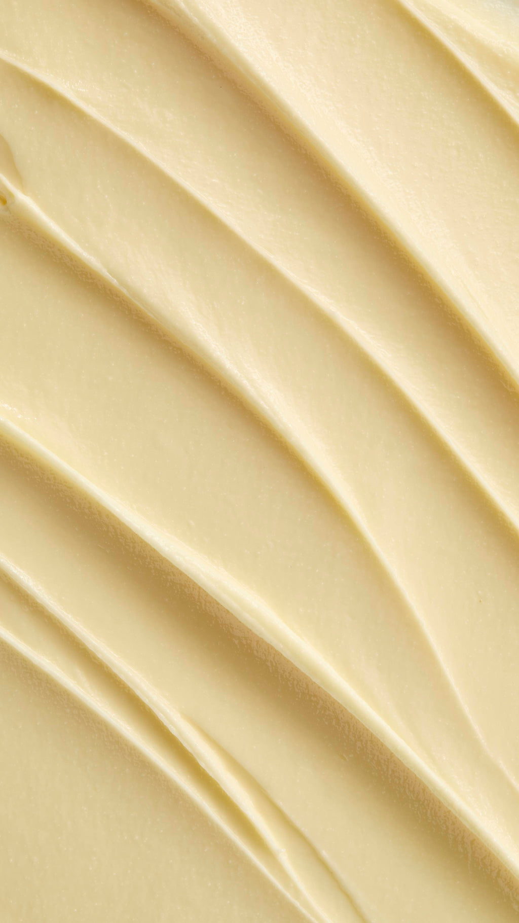 A super close-up image showing diagonal, overlapping smears of the thick, creamy, pale yellow Lush "Oat" facial cleanser. The smooth product has a matte finish and subtle ridged textures.