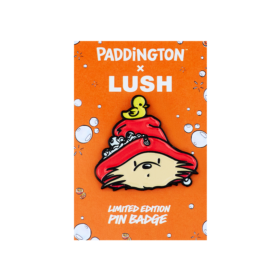 An enamel pin badge from the Paddington x Lush collection. The badge features Paddington Bear’s head wearing a red hat with a small yellow duck on top. It is displayed on an orange backing card with white bubbles and orange slice illustrations.