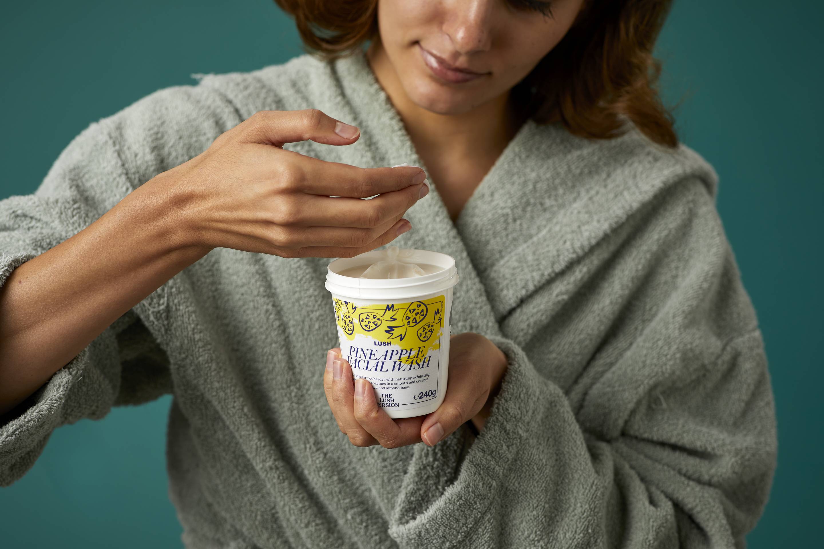 The image shows the model wrapped in a thick, sage-green dressing gown on a teal background. They are holding the white, LUSH version-themed packed pot of the Pineapple facial wash and scooping out the product with their other hand.
