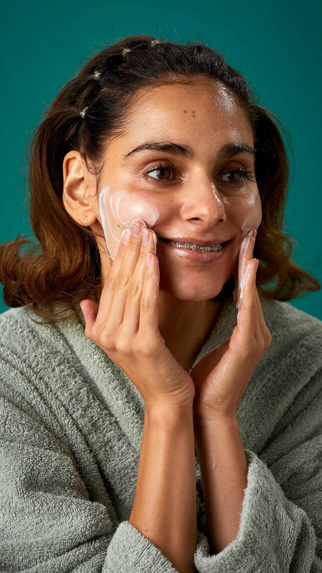The image shows the model wrapped in a thick, sage-green dressing gown on a teal background. They are smiling, showing a glimpse of braces as they apply the Pineapple facial wash over their cheeks.