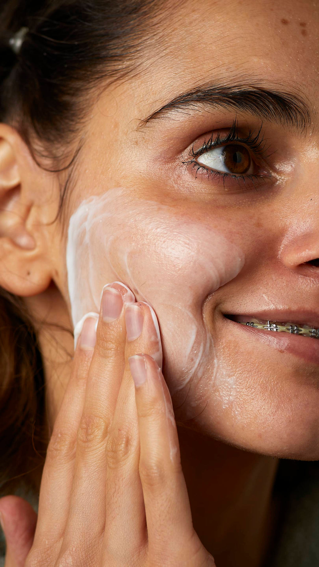 A super close-up image of one of the model's cheeks. They are smiling showing a glimpse of braces as they gently apply the Pineapple facial wash in circular motions over their cheek.