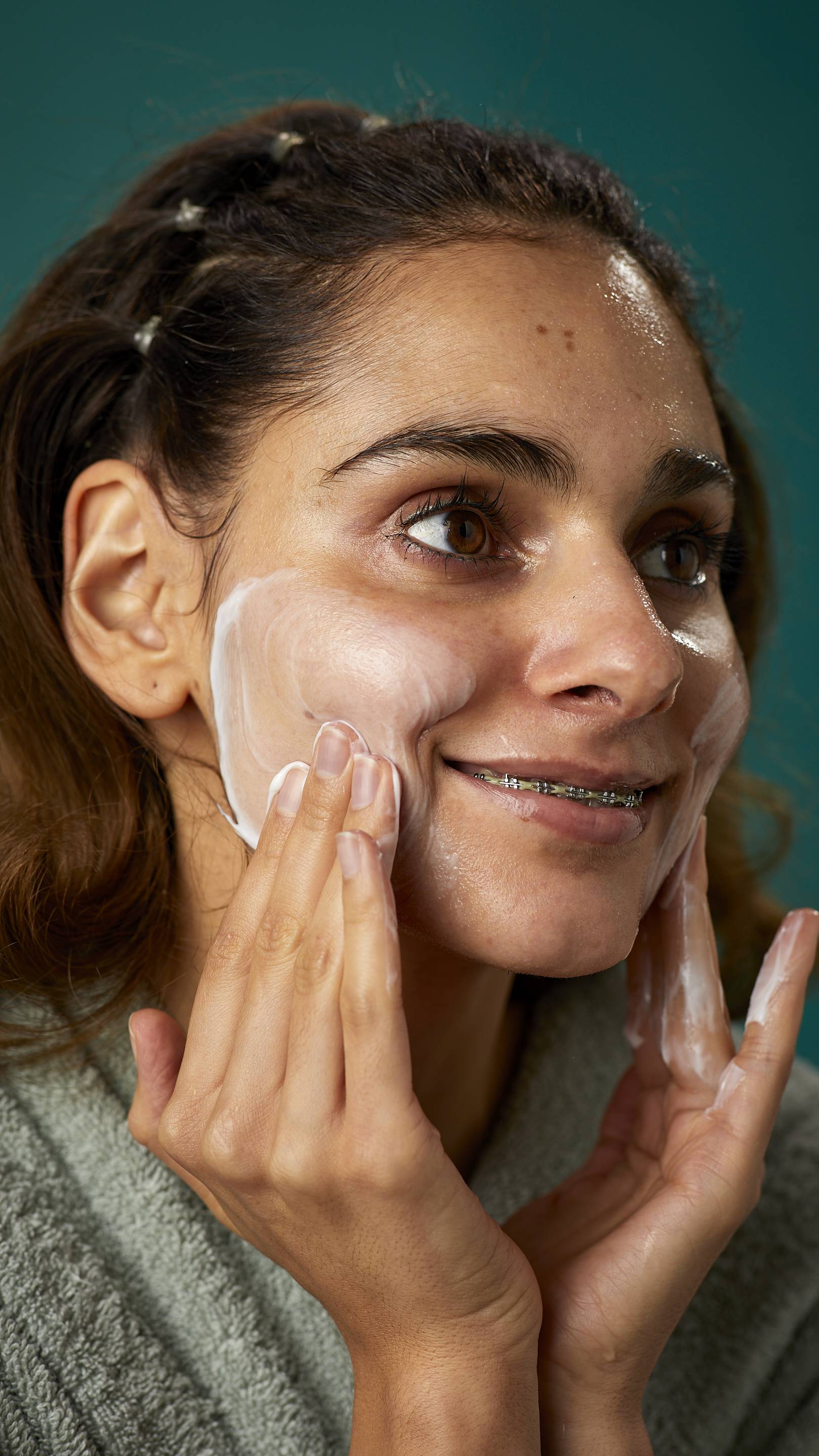 A super close-up image of the model's face. They are smiling showing a glimpse of braces as they gently apply the Pineapple facial wash in circular motions over their cheek.