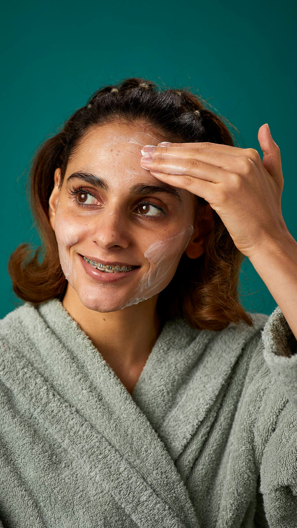 The image shows the model wrapped in a thick, sage-green dressing gown on a teal background. They are smiling, showing a glimpse of braces as they apply the Pineapple facial wash over their cheeks.