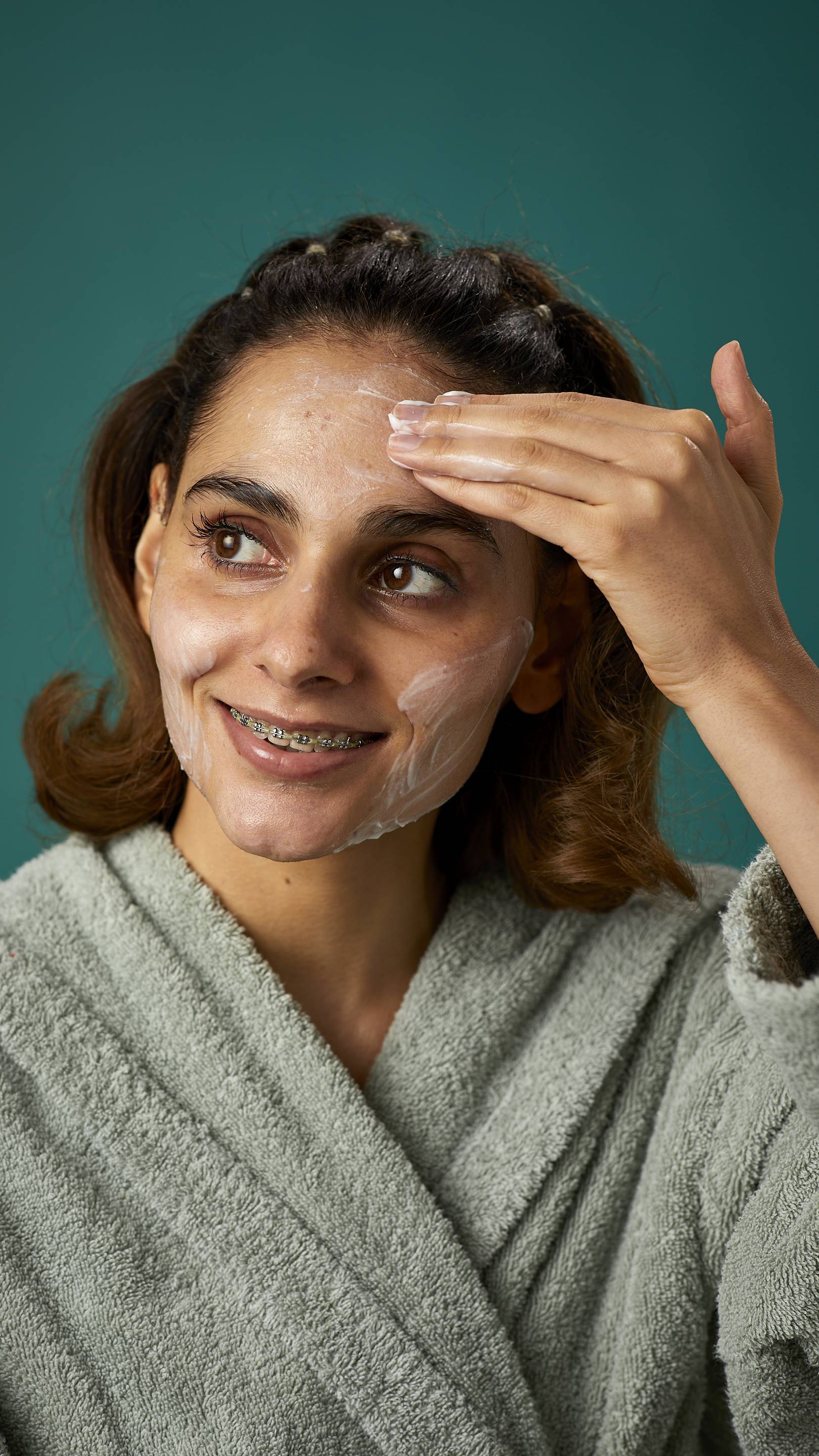 The image shows the model wrapped in a thick, sage-green dressing gown on a teal background. They are smiling, showing a glimpse of braces as they apply the Pineapple facial wash over their cheeks.