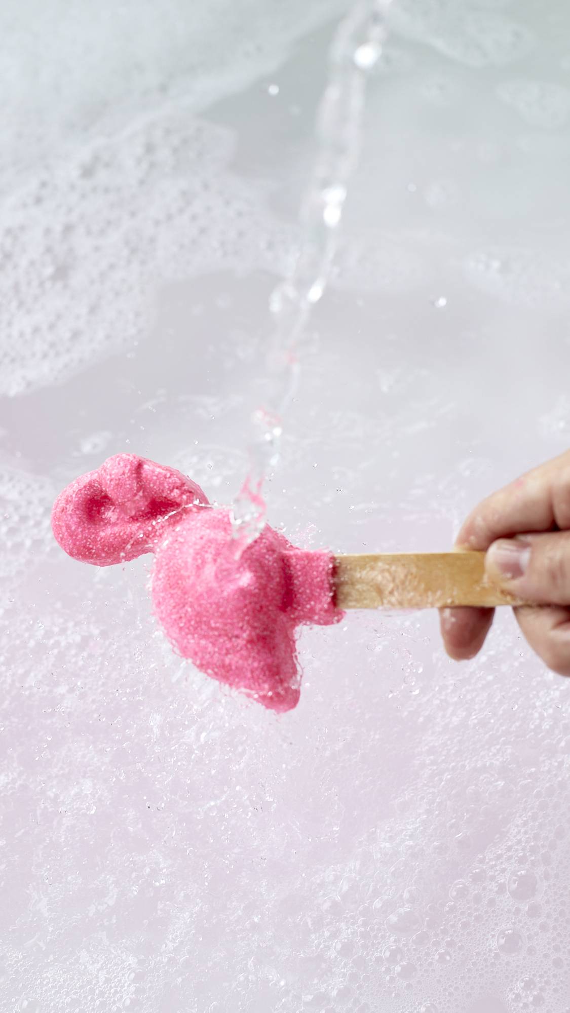 The image shows a close-up shot of the Pink Flamingo reusable bubble bar stick being held under running tap water. The Pink flamingo at the end creates bubbly, delicately rose-tinted water below.