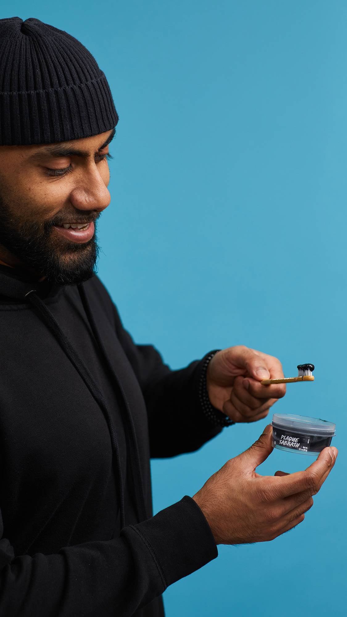 The image shows the model in a black hoodie and hat on a sky-blue background. They are holding an open, clear, recycled plastic pot of Plaque Sabbath toothpaste as they dip the head of their toothbrush into the pot with the other hand.