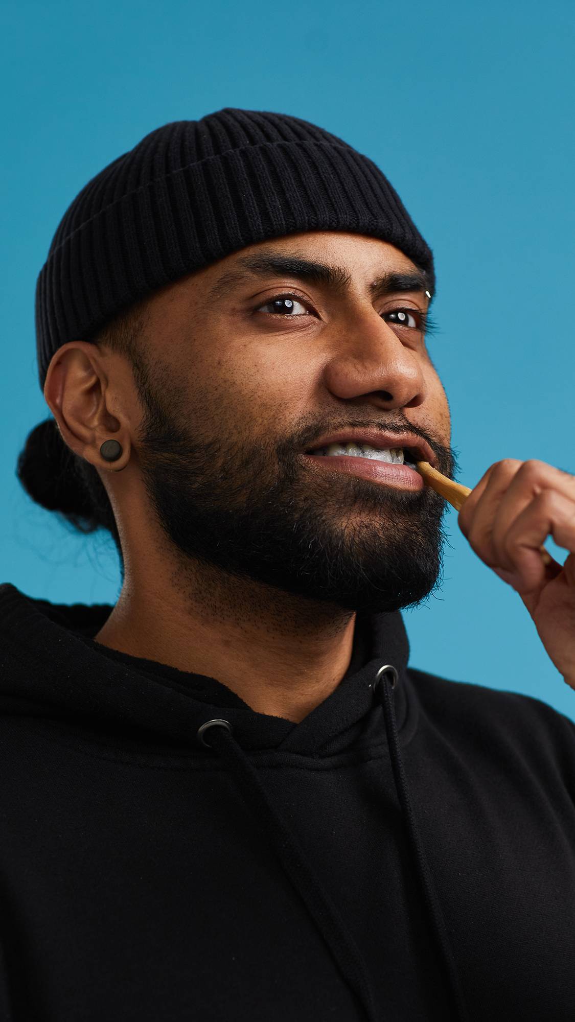 The image shows the model in a black hoodie and hat on a sky-blue background. They are brushing their teeth with a bamboo toothbrush using the Plaque Sabbath toothpaste.