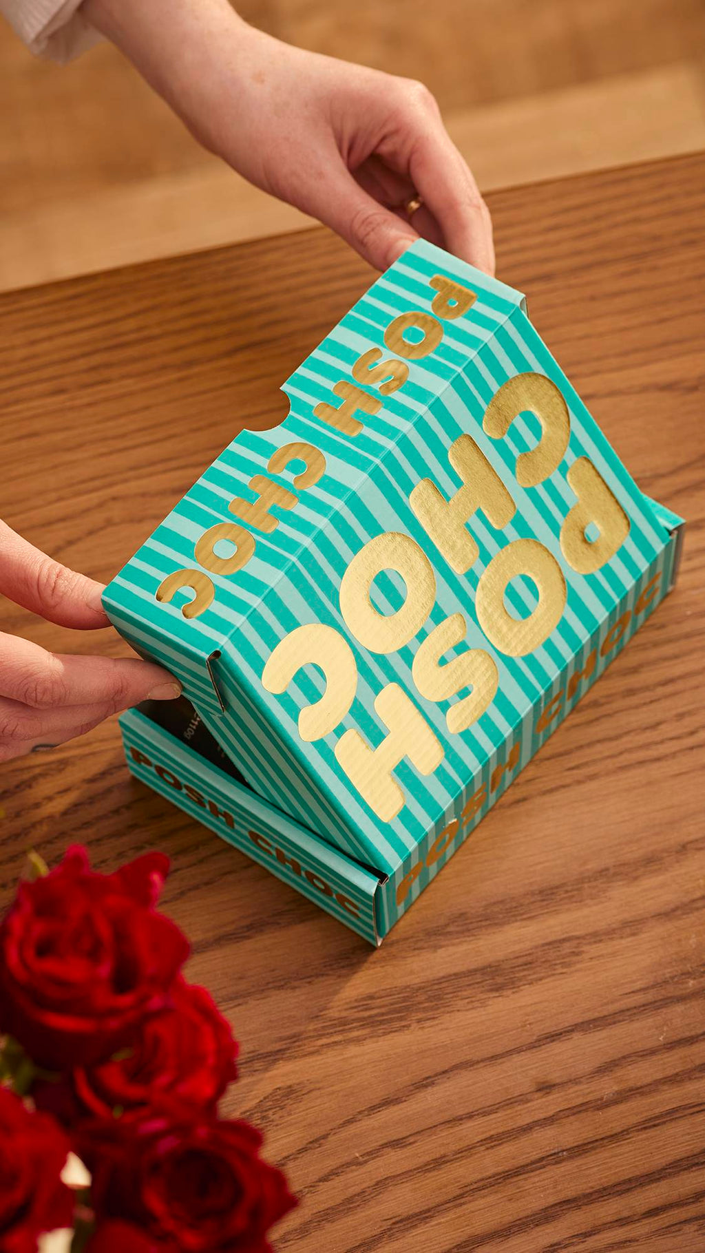 The image shows the Posh Choc, teal-striped gift box on a wooden counter with red roses just visible at the bottom. The model's hand is mid-way through opening the gift box.