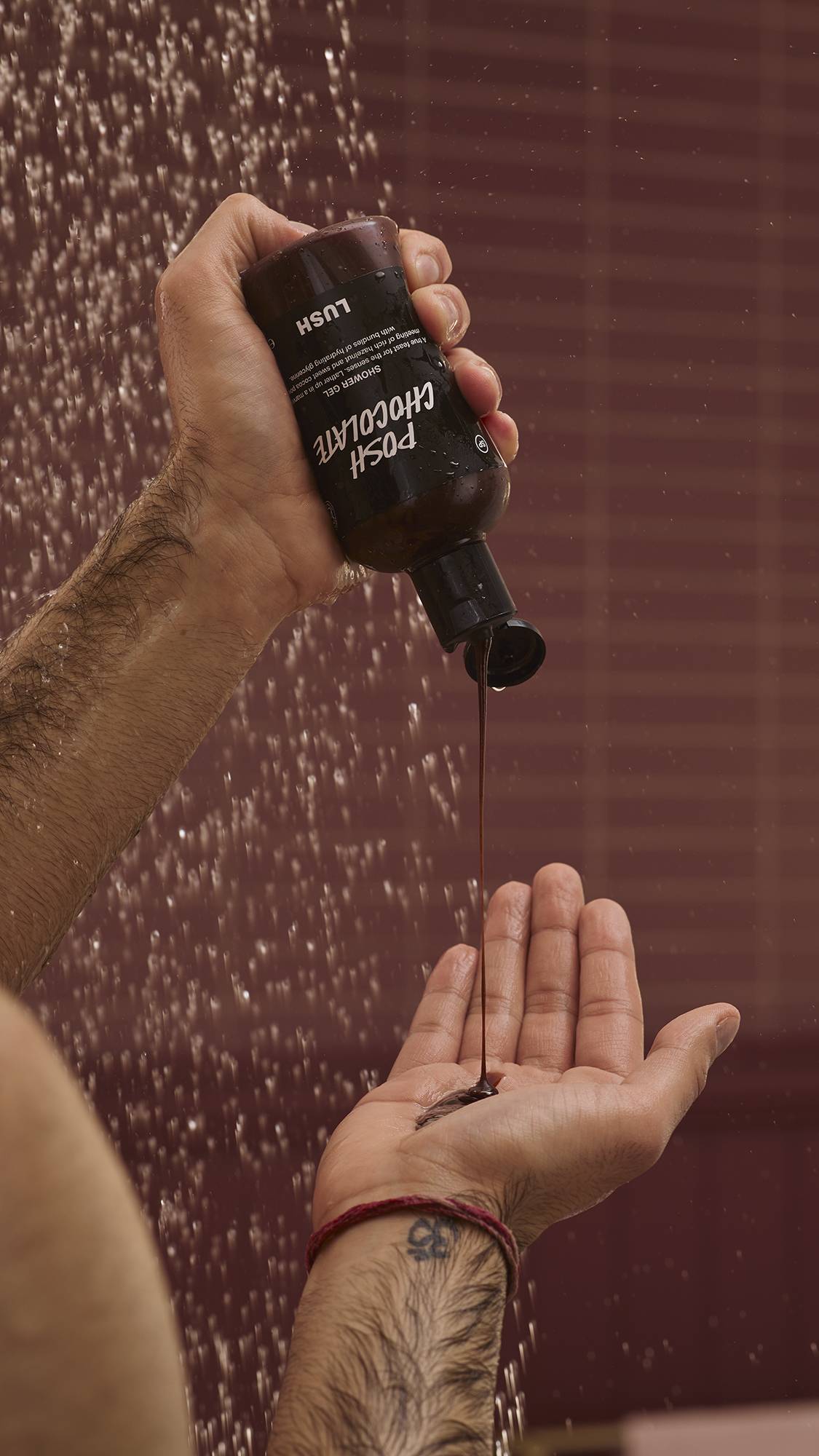 A close-up of the model's hands under running shower water as they squeeze the glossy Posh Chocolate shower gel from the product bottle into their palm. There is a chocolate-coloured background with running shower water.