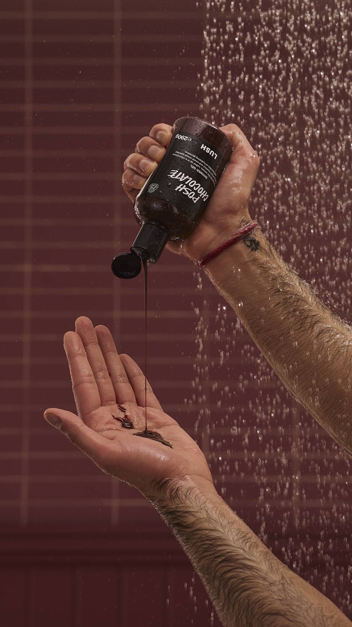 A close-up of the model's hands under running shower water as they squeeze the glossy, molasses-like Posh Chocolate shower gel from the product bottle into their hand. There is a chocolate-coloured background with running shower water.