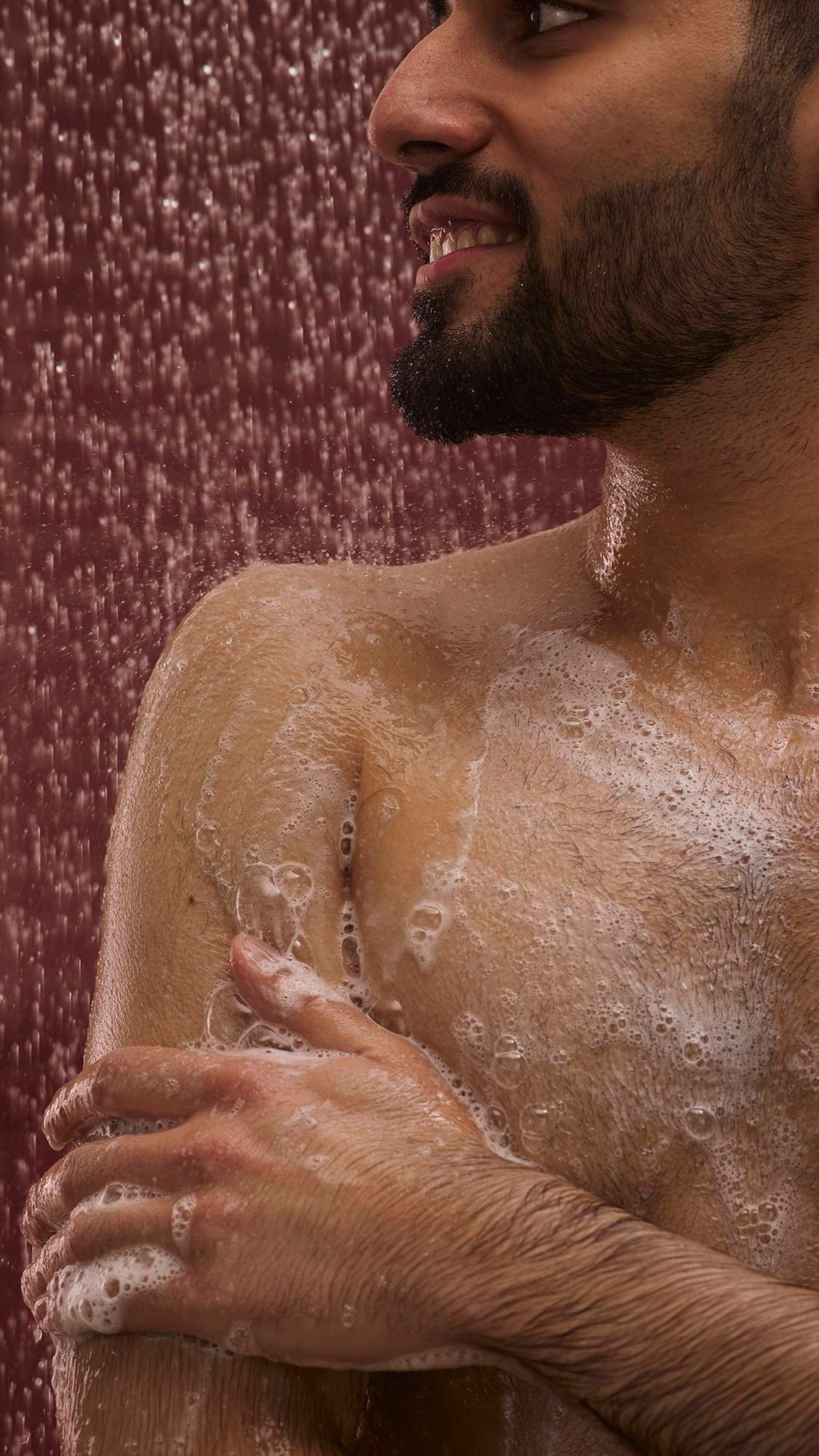 The image shows a dark, chocolate-coloured background with falling shower water. In the foreground, the model stands facing the camera as they lather up their shoulder with the glossy Posh Chocolate shower gel to create a soft foam.