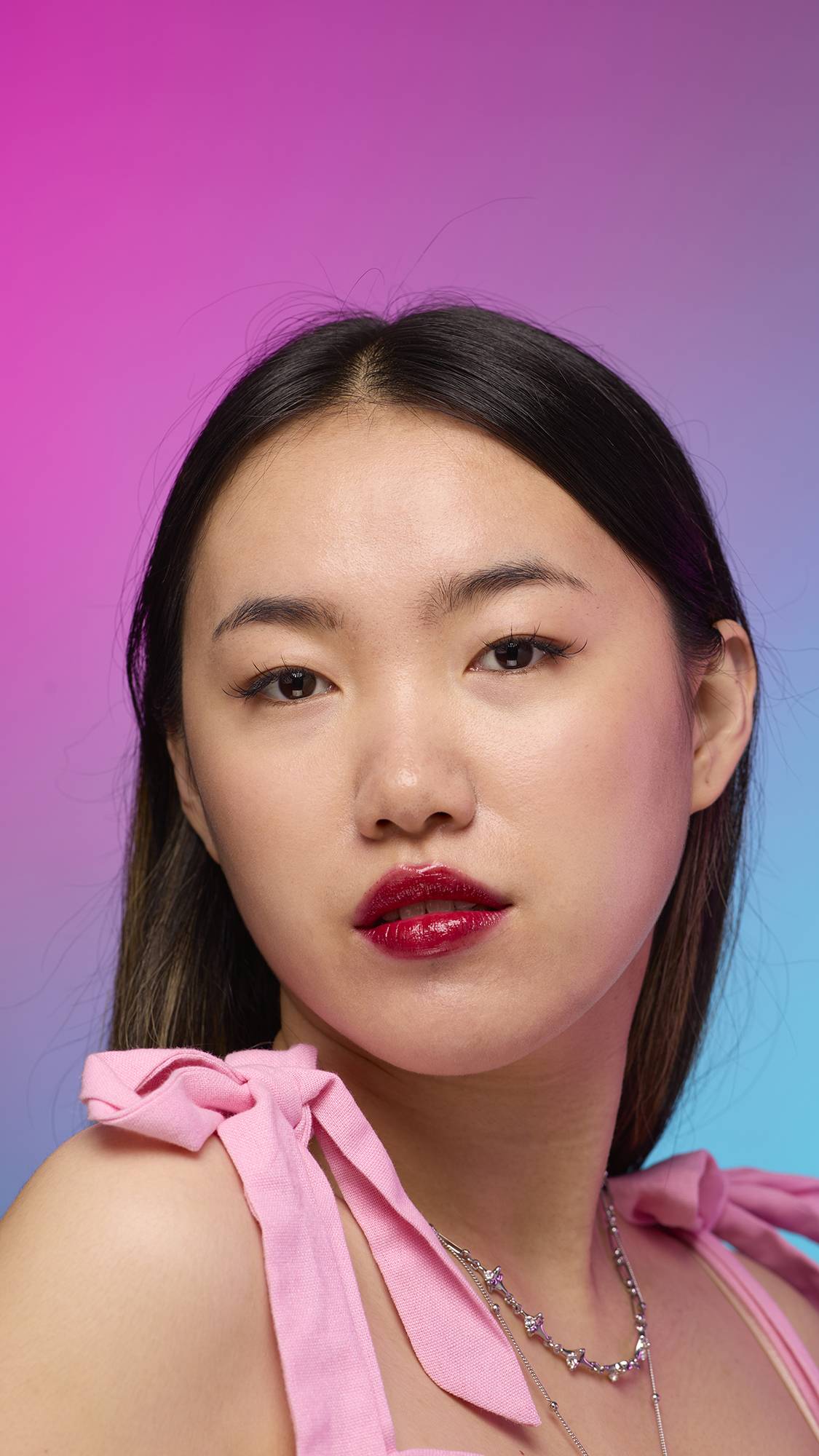The model has long, straight dark hair and is on a pastel pink, blue and purple hue background. Their make-up has been done with the focal point being the juicy coating of carmine-red tint on their lips created by the Red Maple lip jelly.