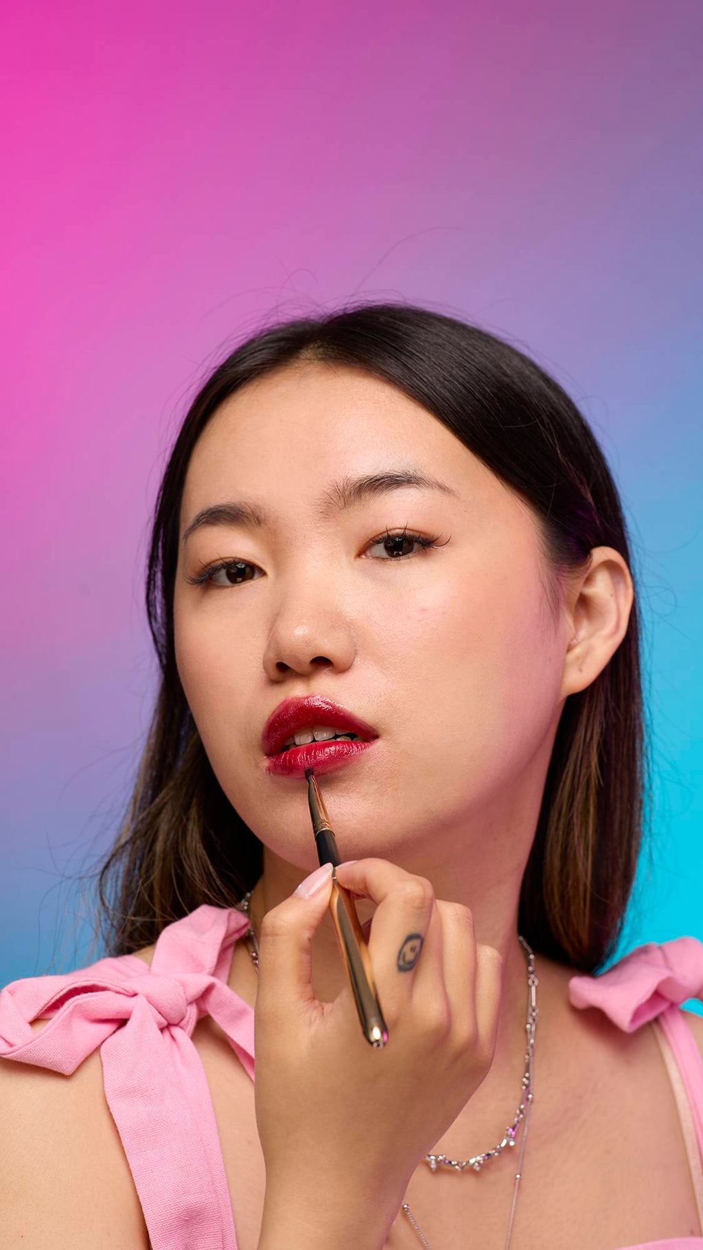 The model has long, dark hair and is on a pastel pink, blue and purple hue background. They are holding a thin, long brush to their face as they apply the vivid, Red Maple lip jelly.