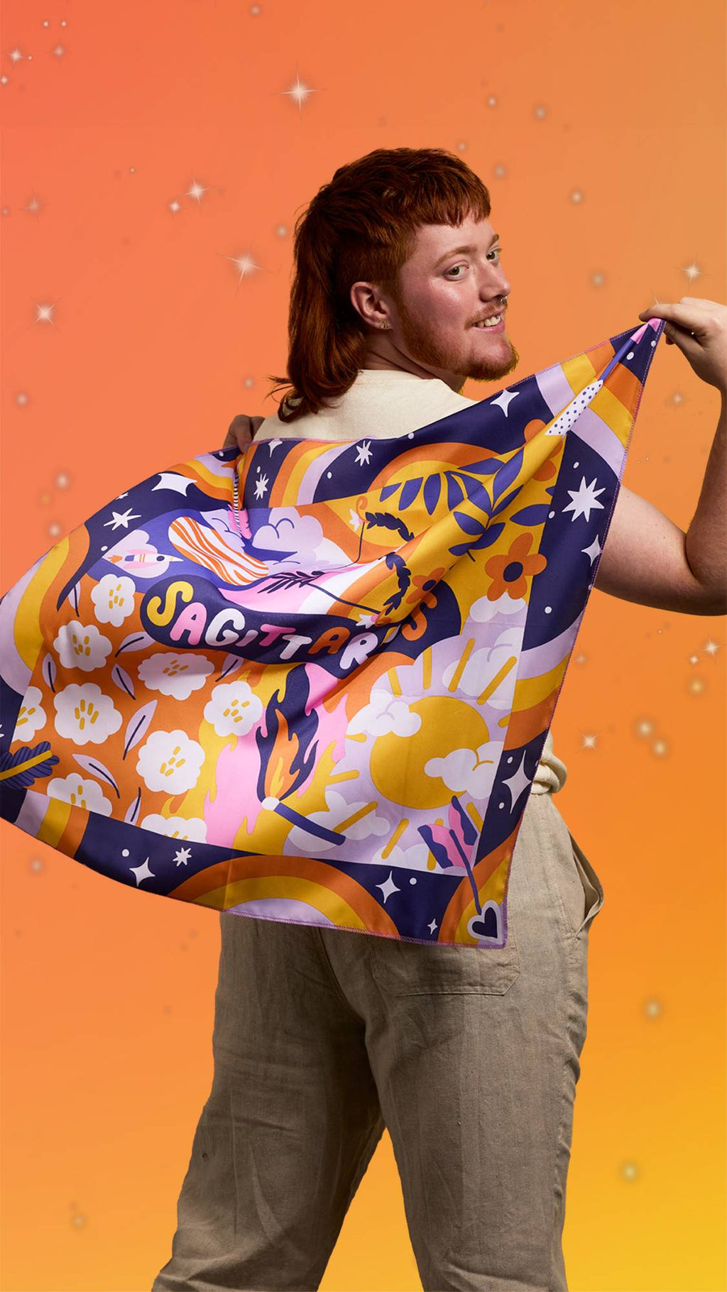 The image shows a yellow and orange blurred background scattered with sparkles. A model is standing, facing away as they hold the Sagittarius knot wrap over their shoulders, almost like a cape, to show the design off.