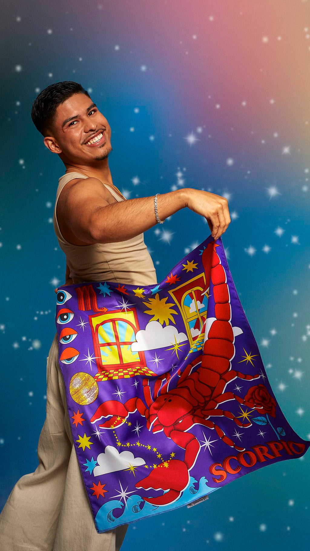 The image shows the model wearing a beige tank top and linen trousers. They are smiling on a pastel, sparkling background and holding the Scorpio knot wrap behind their back showing the full design.