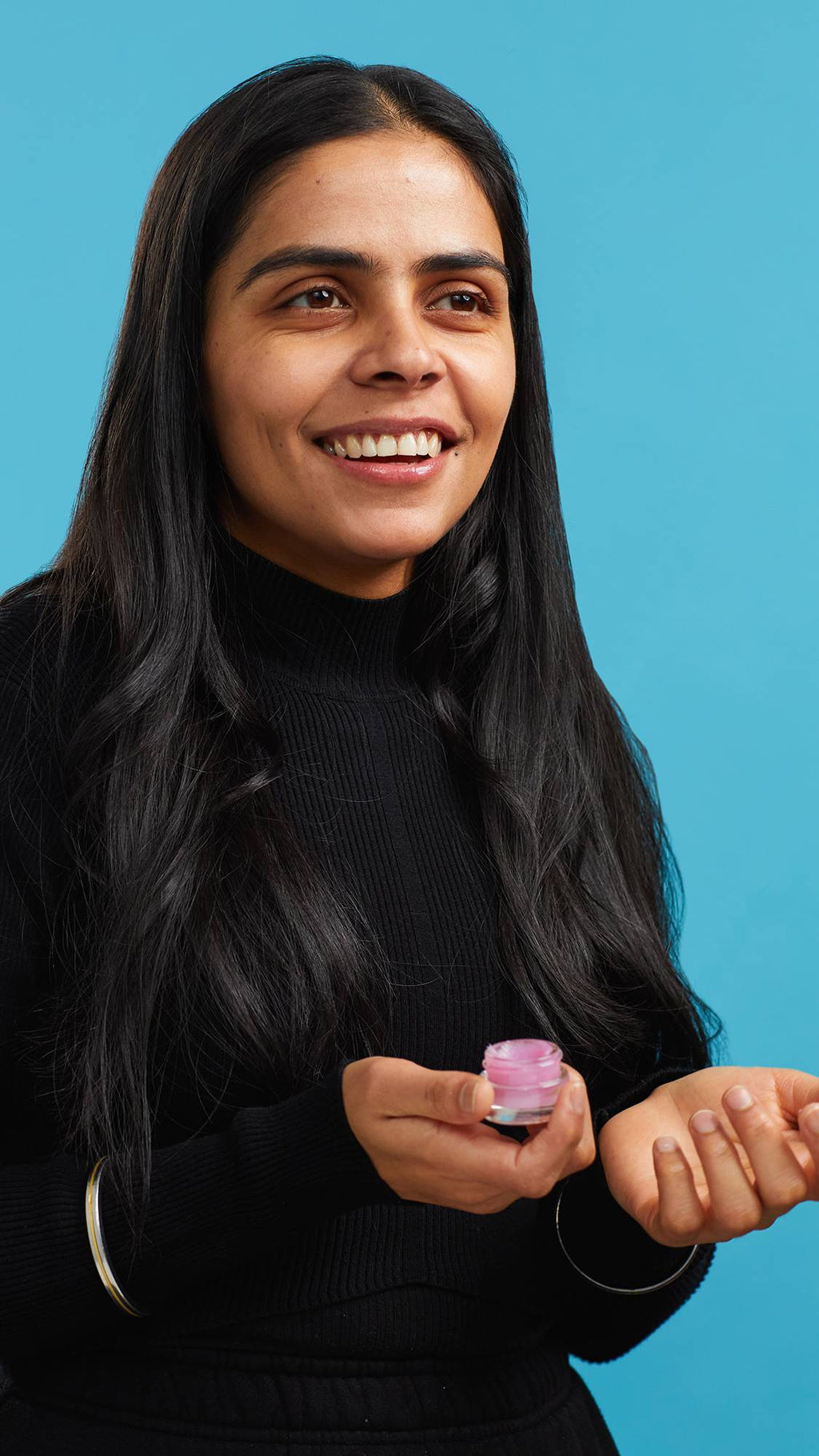 A close-up shot of the model on a sky-blue background wearing a black turtleneck. They are holding a small, glass pot of the pink Sex Bomb and smiling to reveal juicy, glossy lips.
