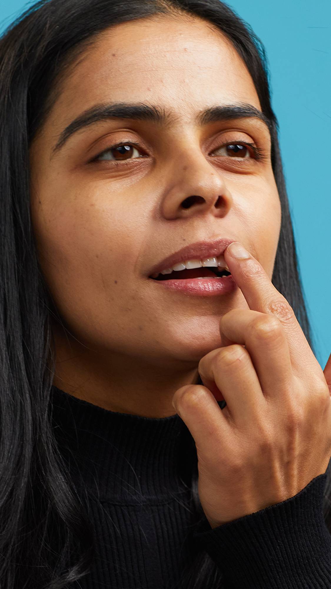 A close-up shot of the model on a sky-blue background wearing a black turtleneck. They are holding a small, glass pot of the pink Sex Bomb lip gloss as they apply it to their lips.