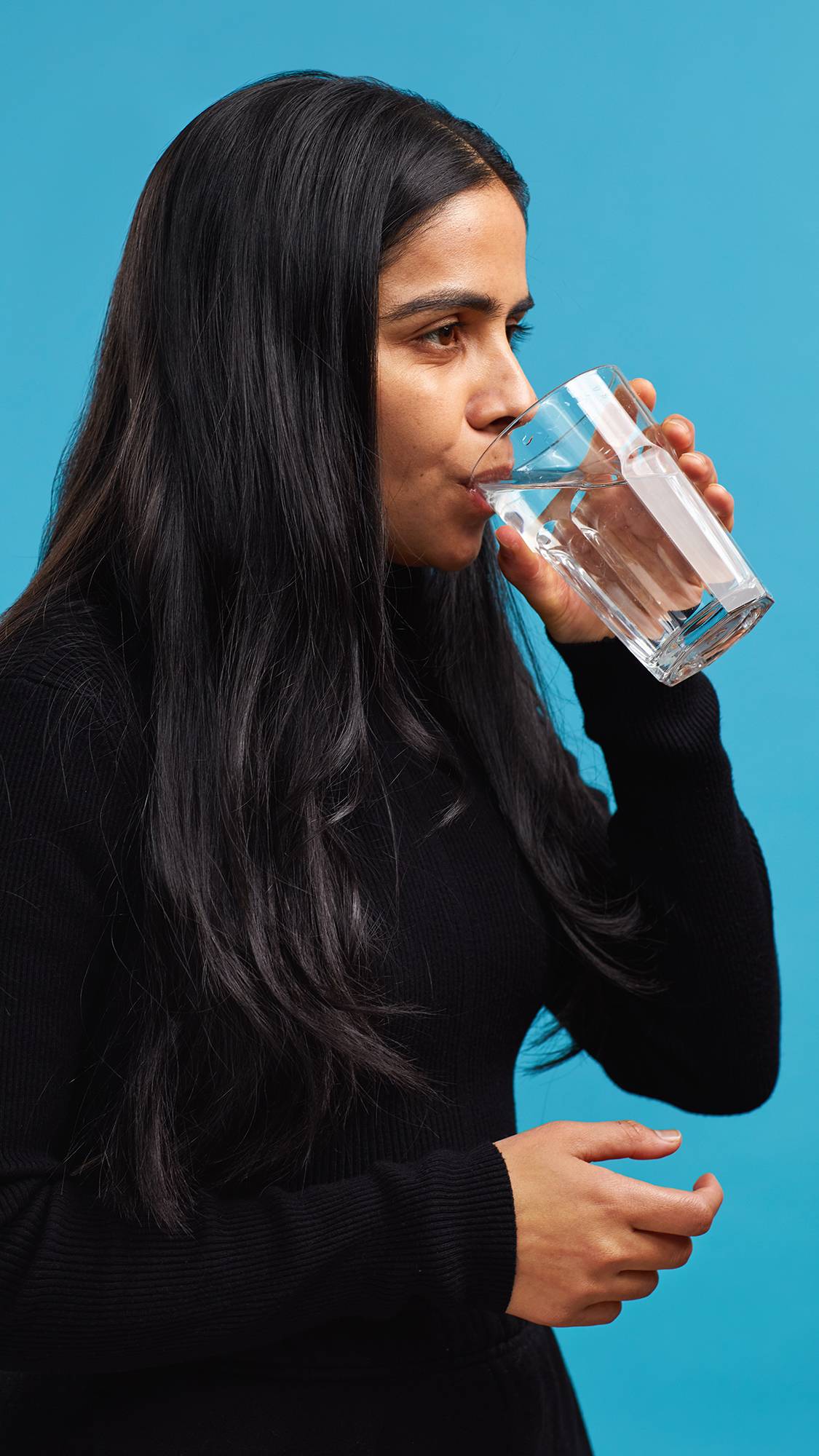 The image shows the model wearing a black turtleneck on a sky-blue background. They have just placed one of the Shimmy Shimmy mouthwash tabs into their mouth and are taking a sip of water from a glass.