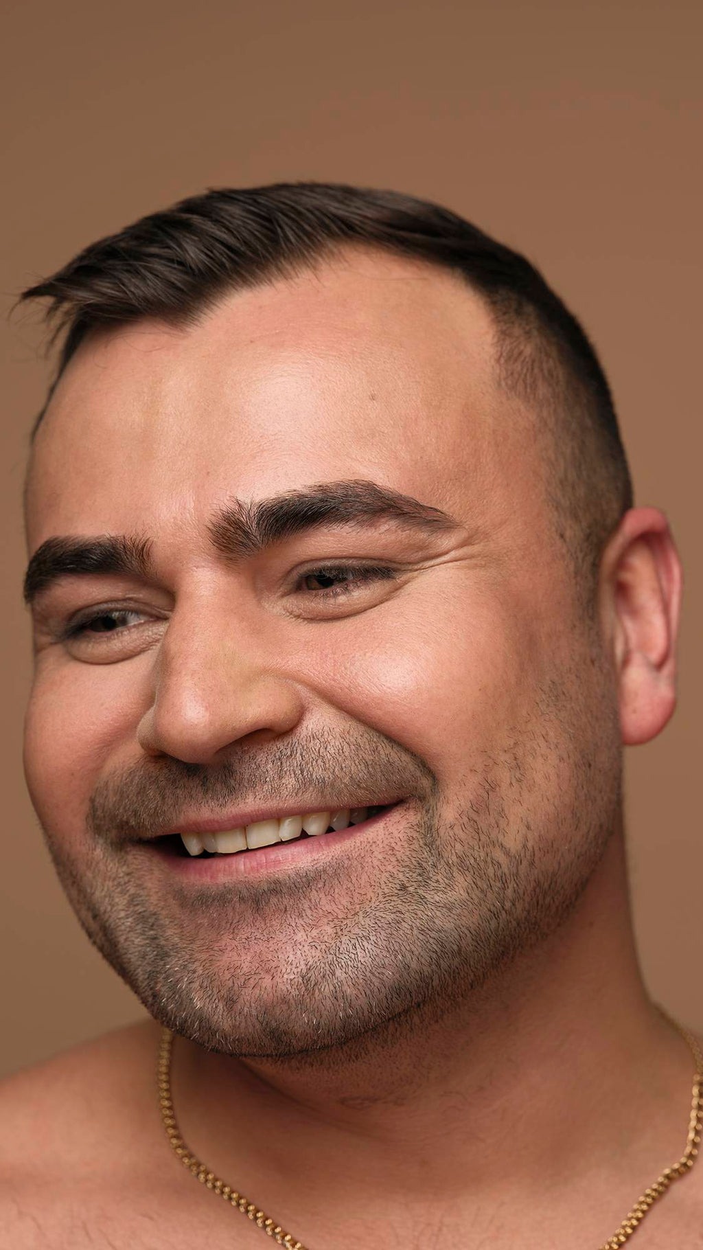 A close-up side profile of a smiling person with short dark hair and facial stubble, wearing a gold chain and looking off-camera against a neutral background.