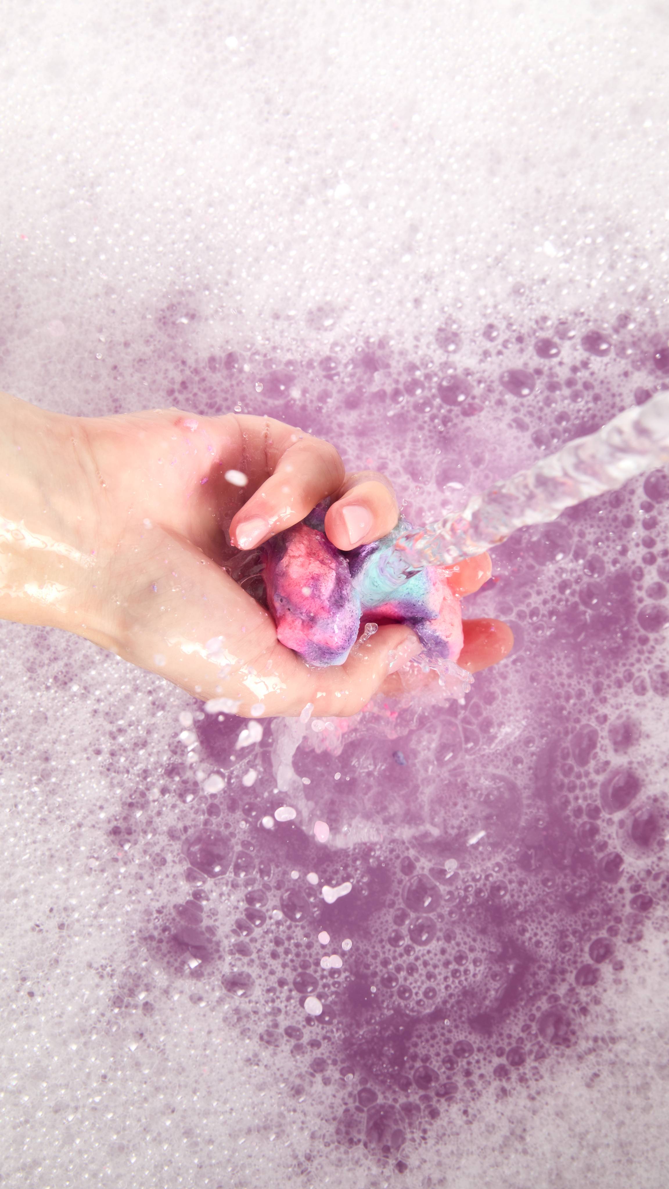 The image shows the model's hand as it crumbles a mini Sleepy bubble bar under running tap water to produce a mountain of bubbles below and delicate, purple bath water.