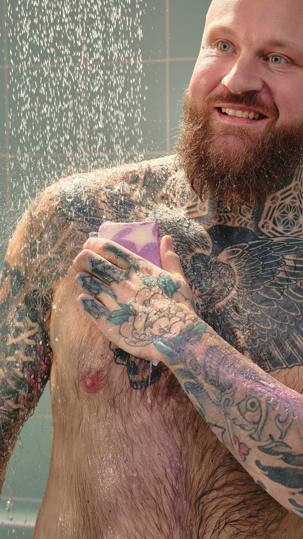 A smiling man with chest and arm tattoos uses a purple Sleepy soap bar under a shower spray. The soap features a white star design, and the background shows light green tiles.