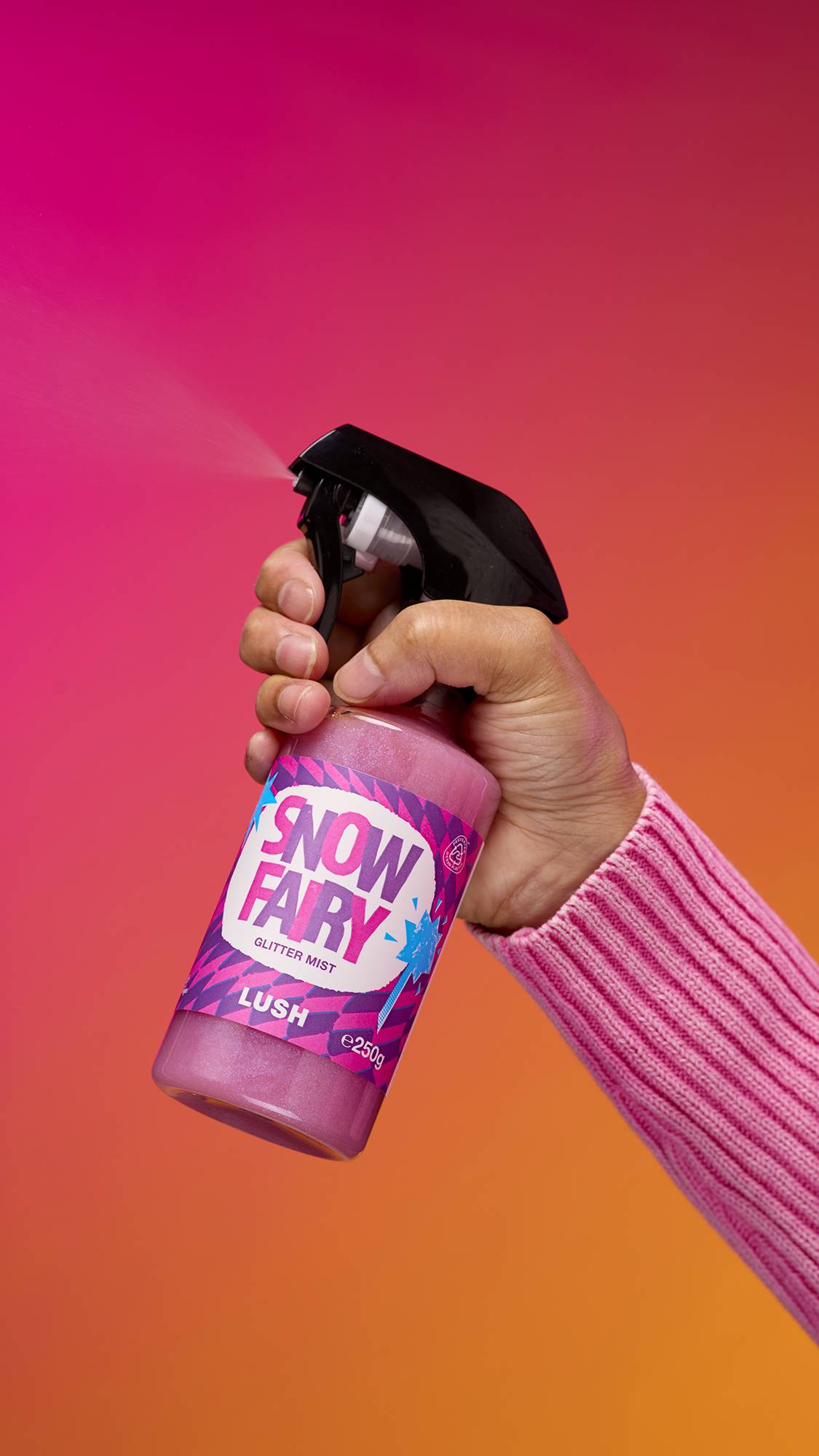 An arm is reaching from the right-hand side. The hand is spritzing a transparent, cylindrical plastic spray bottle with pink, shimmering liquid and a black top. A colourful label ready "Snow Fair Glitter Mist" on a pink-to-orange gradient background.