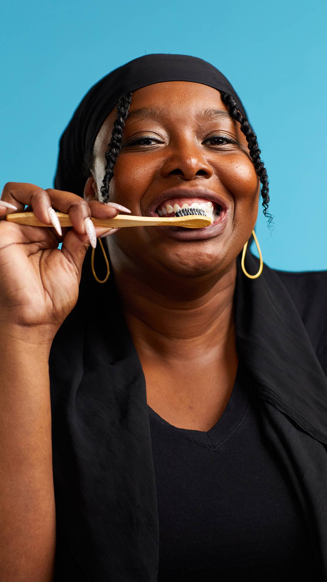 The image shows the model in a black top on a sky-blue background. They are brushing their teeth with a bamboo toothbrush using the Soother toothpaste and smiling.