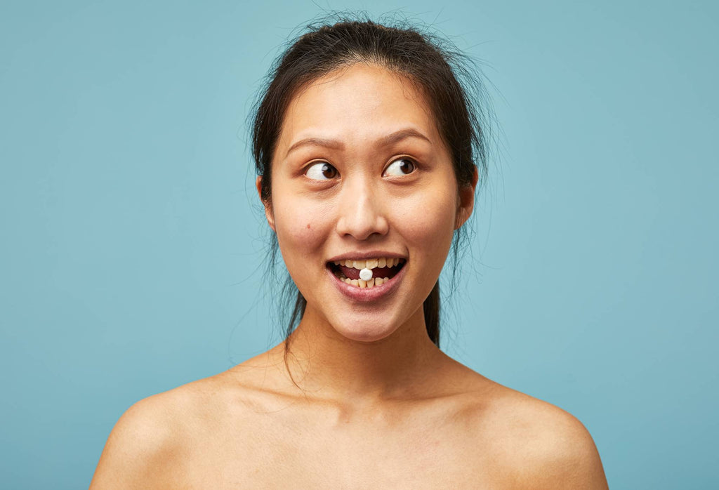 A person shows off their teeth and holds a small white Sparkle toothpaste tab between their front teeth.