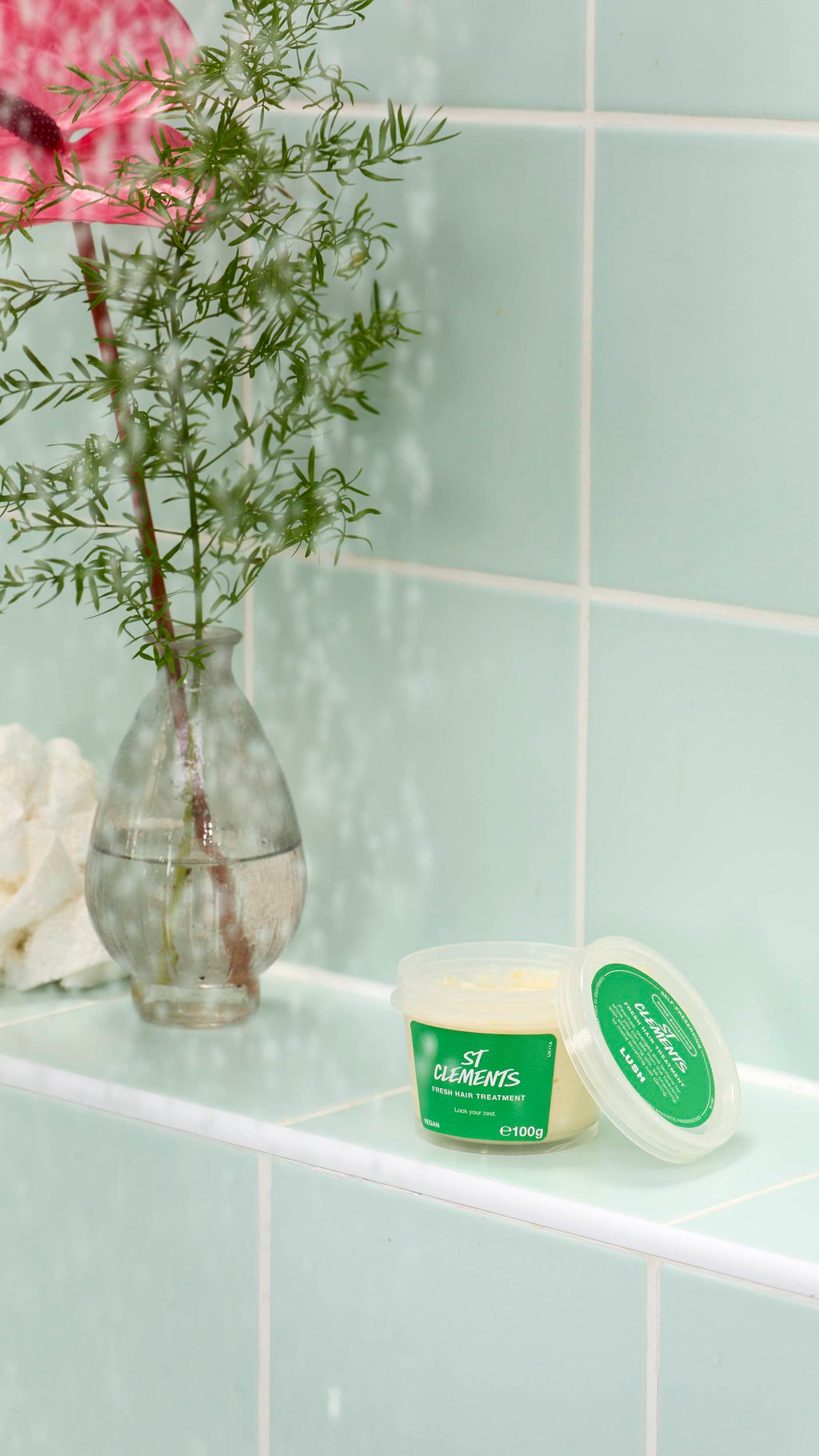 An open pot of pale yellow "St Clements" fresh hair treatment by Lush sits on a light green tiled ledge. In the background, a glass vase holds a green plant and a pink flower, partially obscured by falling water droplets.