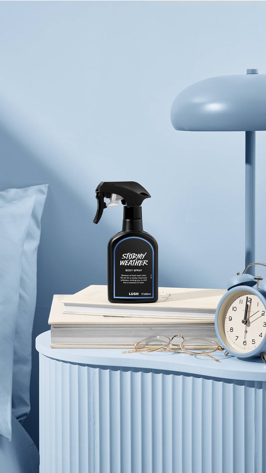 The image shows a recycled black plastic LUSH spray bottle of "Stormy Weather" sitting on a pile of books on a sea-blue bedside table. A matching lamp, alarm clock, and glasses sit nearby, next to a blue bed and wall.