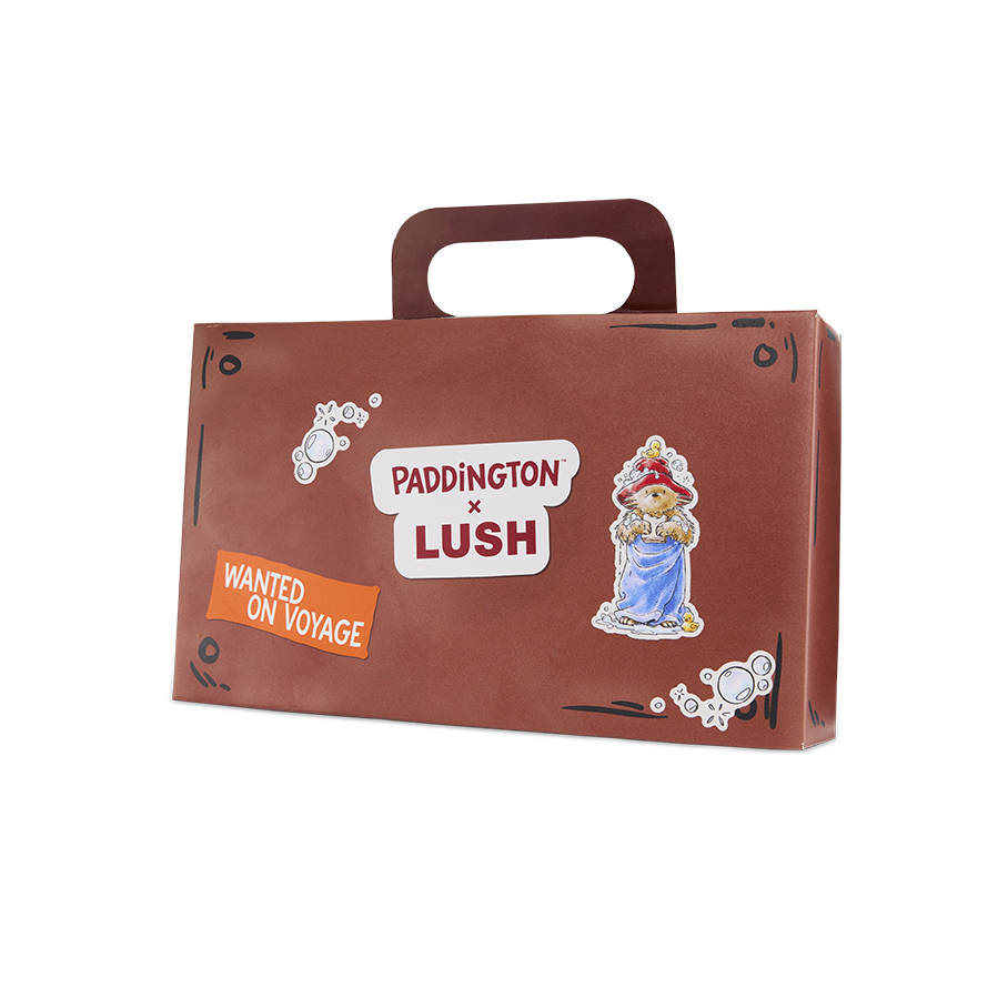 Suitcase. A brown cardboard gift box shaped like a suitcase with a carry handle. The front features 'PADDINGTON x LUSH' branding, an illustration of Paddington Bear in a towel, and a 'WANTED ON VOYAGE' sticker.