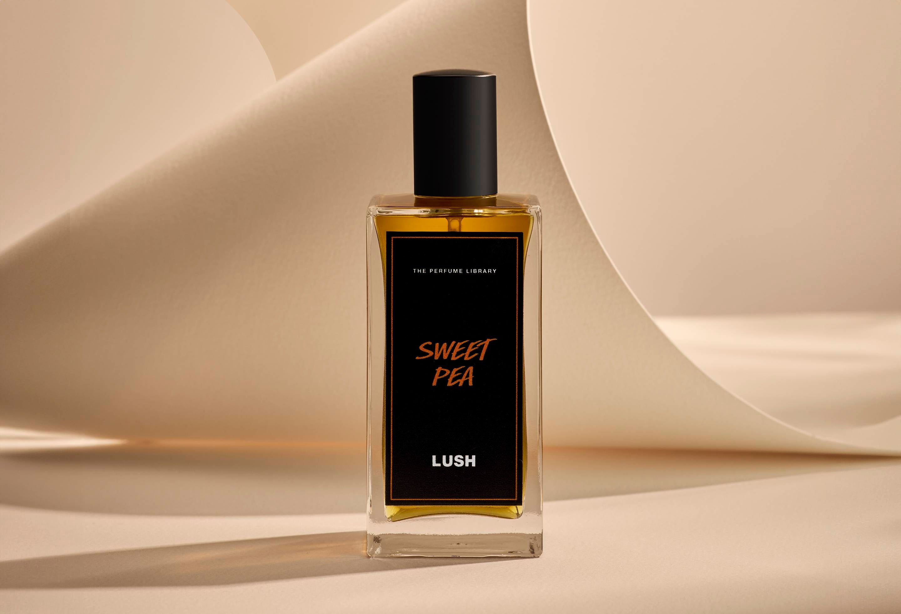 The image shows the Sweet Pea, glass perfume bottle. It is sitting on a soft, beige background with curved, cream paper creating soft, gliding shapes behind the product.