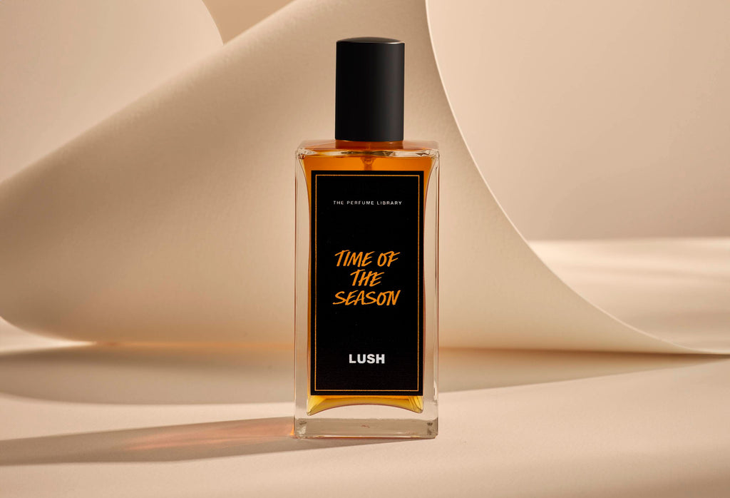 The image shows the Time Of The Season glass perfume bottle. It is sitting on a soft, beige background with curved, cream paper creating soft, gliding shapes behind the product.