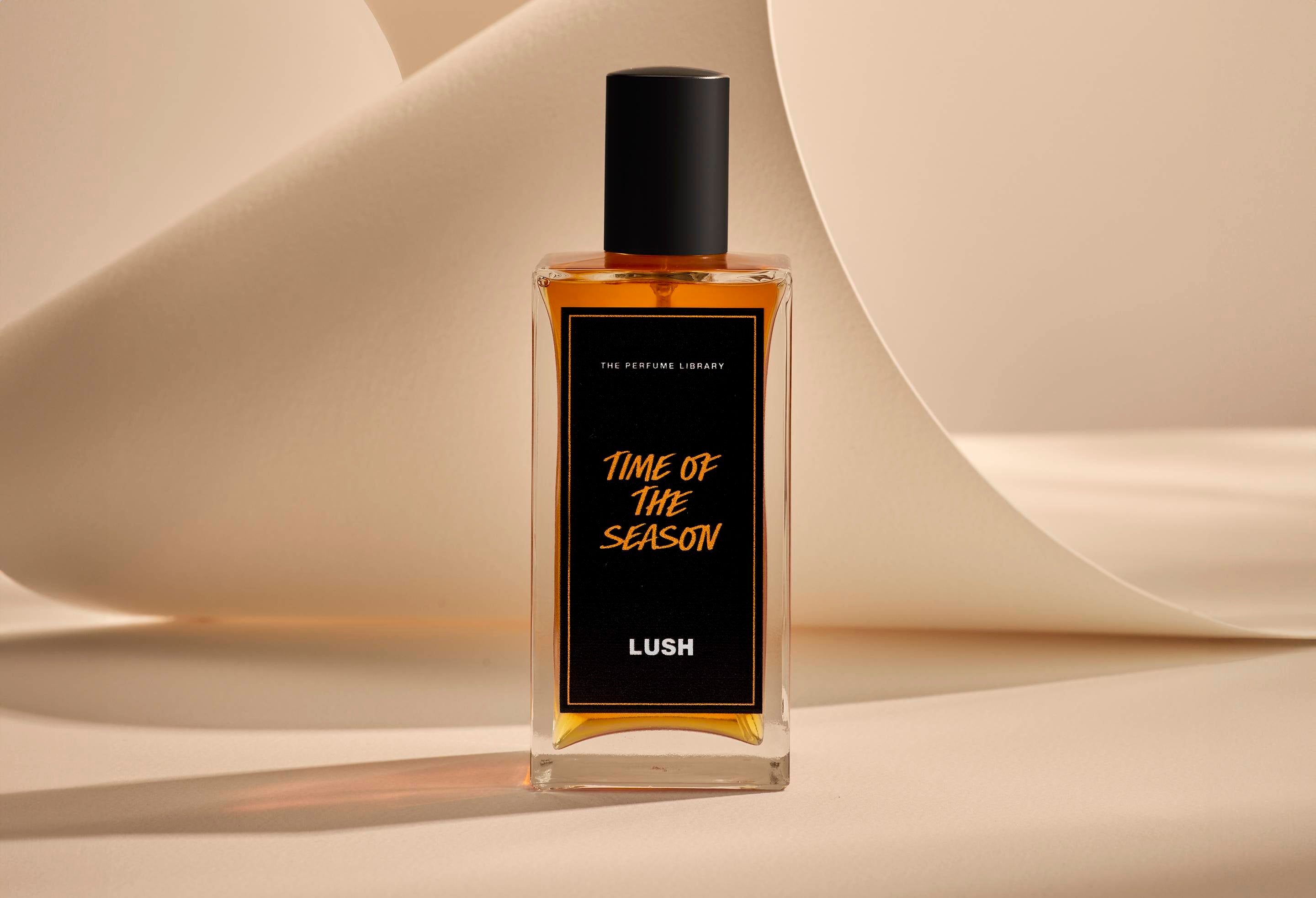 The image shows the Time Of The Season glass perfume bottle. It is sitting on a soft, beige background with curved, cream paper creating soft, gliding shapes behind the product.