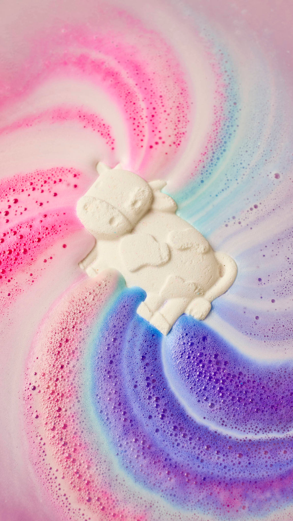 An aerial view of the cow-shaped Toby's Magic Cow as it is dissolved in the bath. The product slowly dissolves as it gives off rippling swirls of velvety foam with streaks of pink, blue and purple.