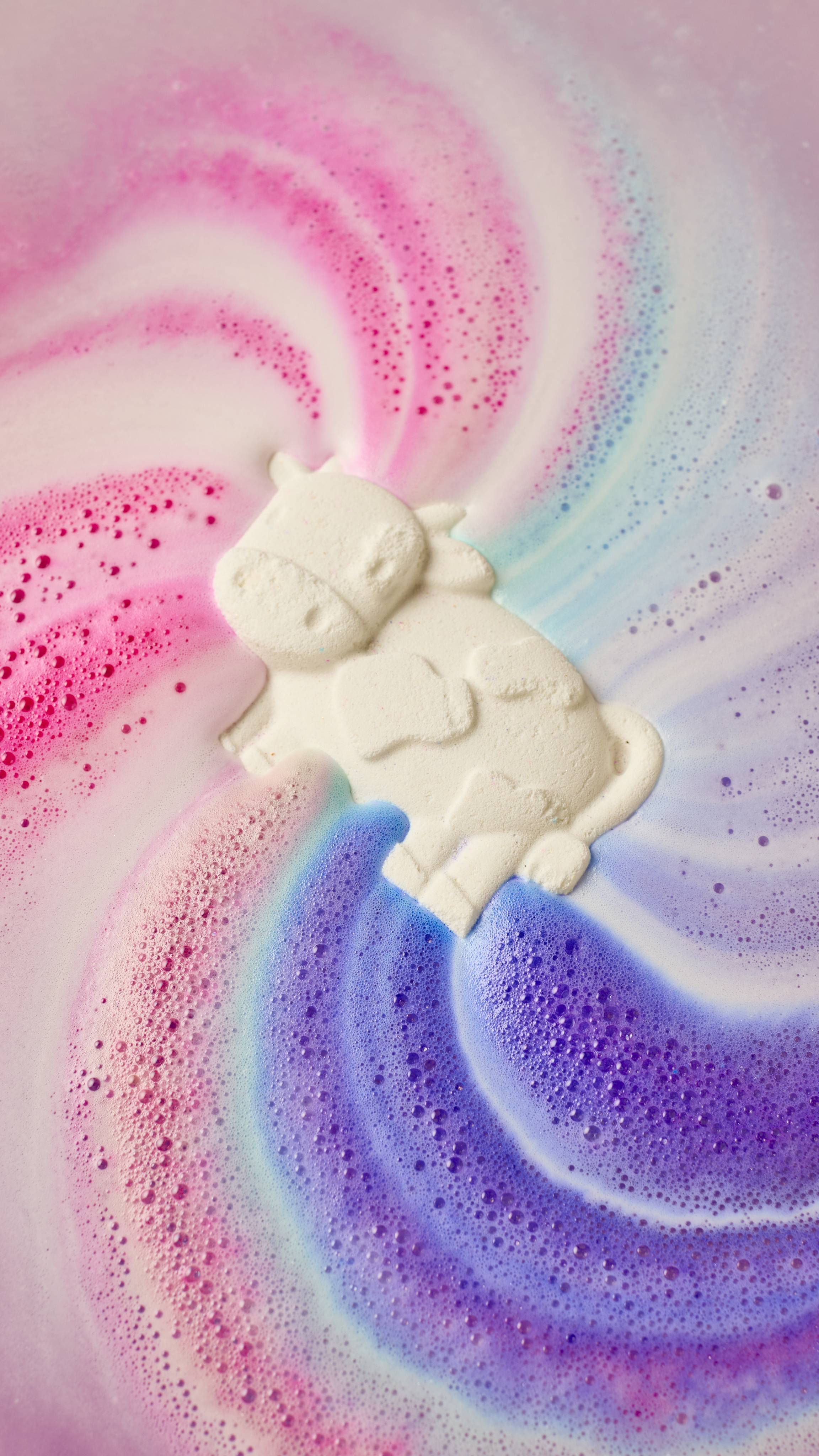 An aerial view of the cow-shaped Toby's Magic Cow as it is dissolved in the bath. The product slowly dissolves as it gives off rippling swirls of velvety foam with streaks of pink, blue and purple.