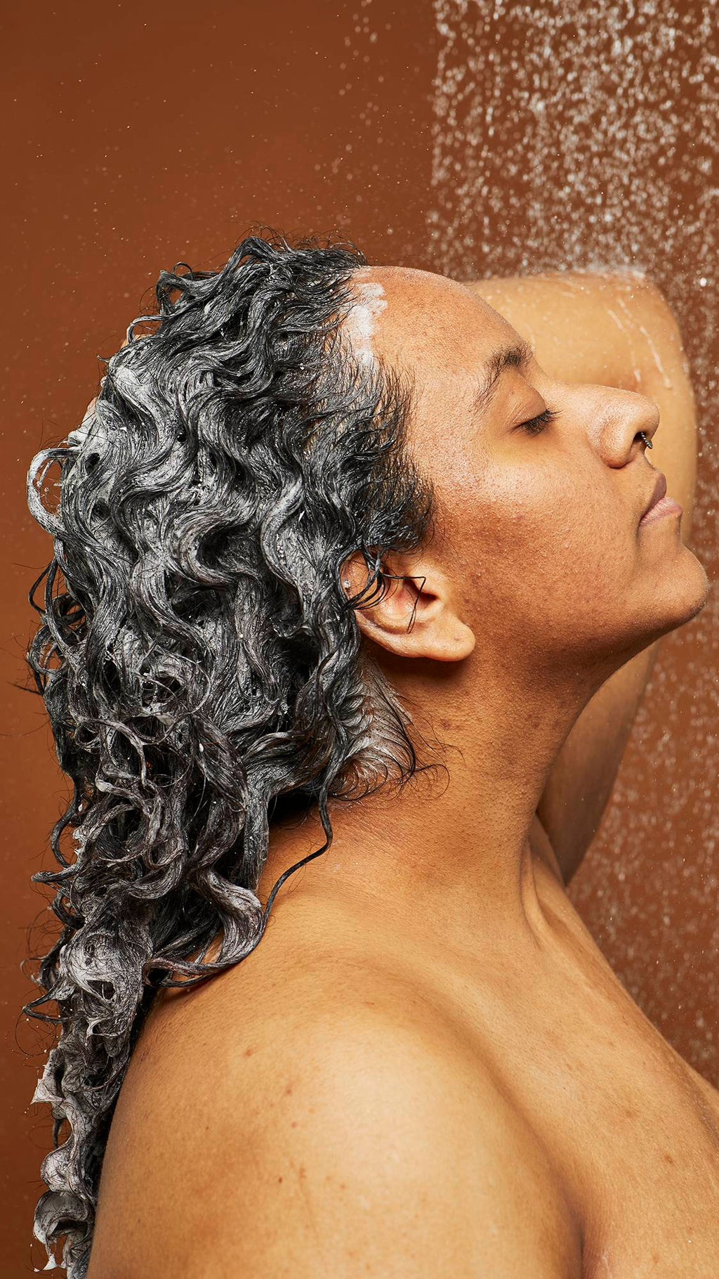 Model is standing under shower water lathering up the shampoo as it falls down their back on a deep earth-tone background.