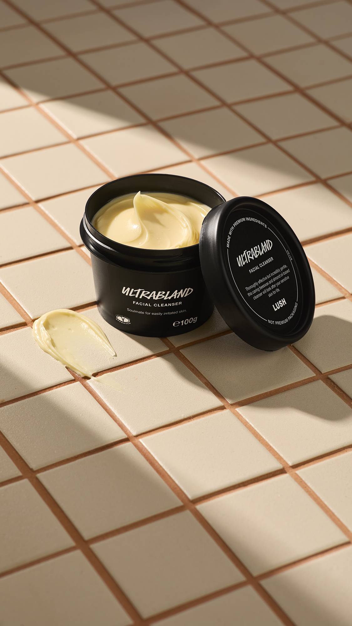 A black pot of Lush Ultrabland Facial Cleanser sits on a beige tiled surface with the lid leaning against it and a smear of the thick yellow cream nearby.