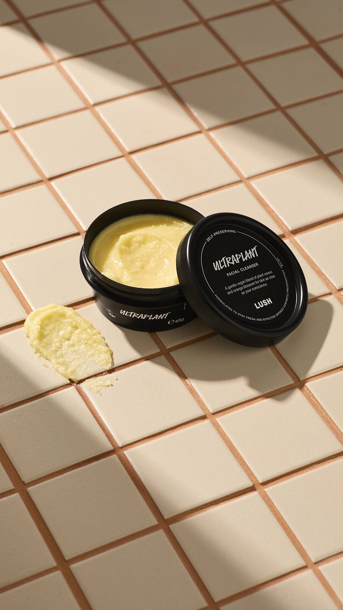 An open black tub of Lush 'Ultraplant' facial cleanser sits on a beige tiled surface. Beside it is a small smear of the thick, pale yellow product. The lid rests against the pot, showing the product name and Lush logo in white text.