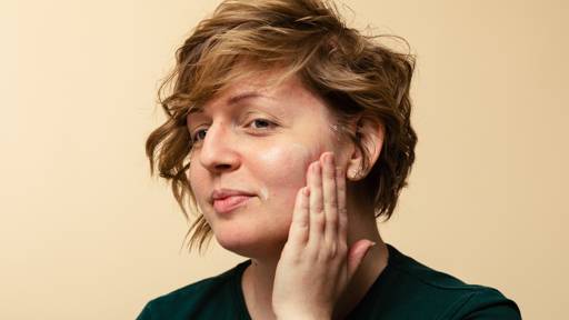 A person with short, wavy hair smiles softly as they rub Vanishing Cream, a light moisturiser, into their cheek with their hand.
