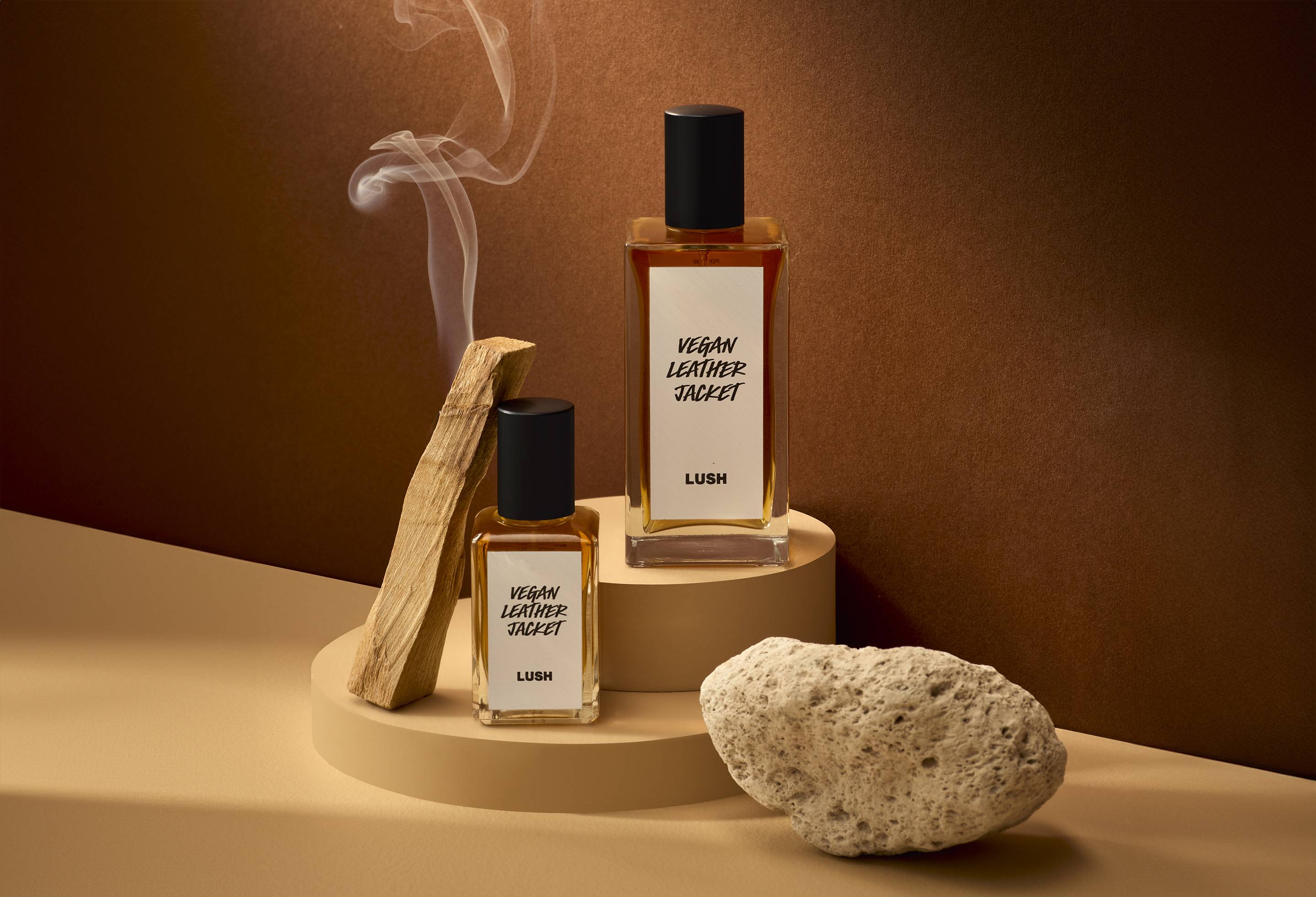 The image shows a warm, bark-brown background. In the foreground are the two different-sized bottles of the Vegen Leather Jacket perfume on a small, stacked pedestal with a smoking piece of wood and a porous rock beside them.