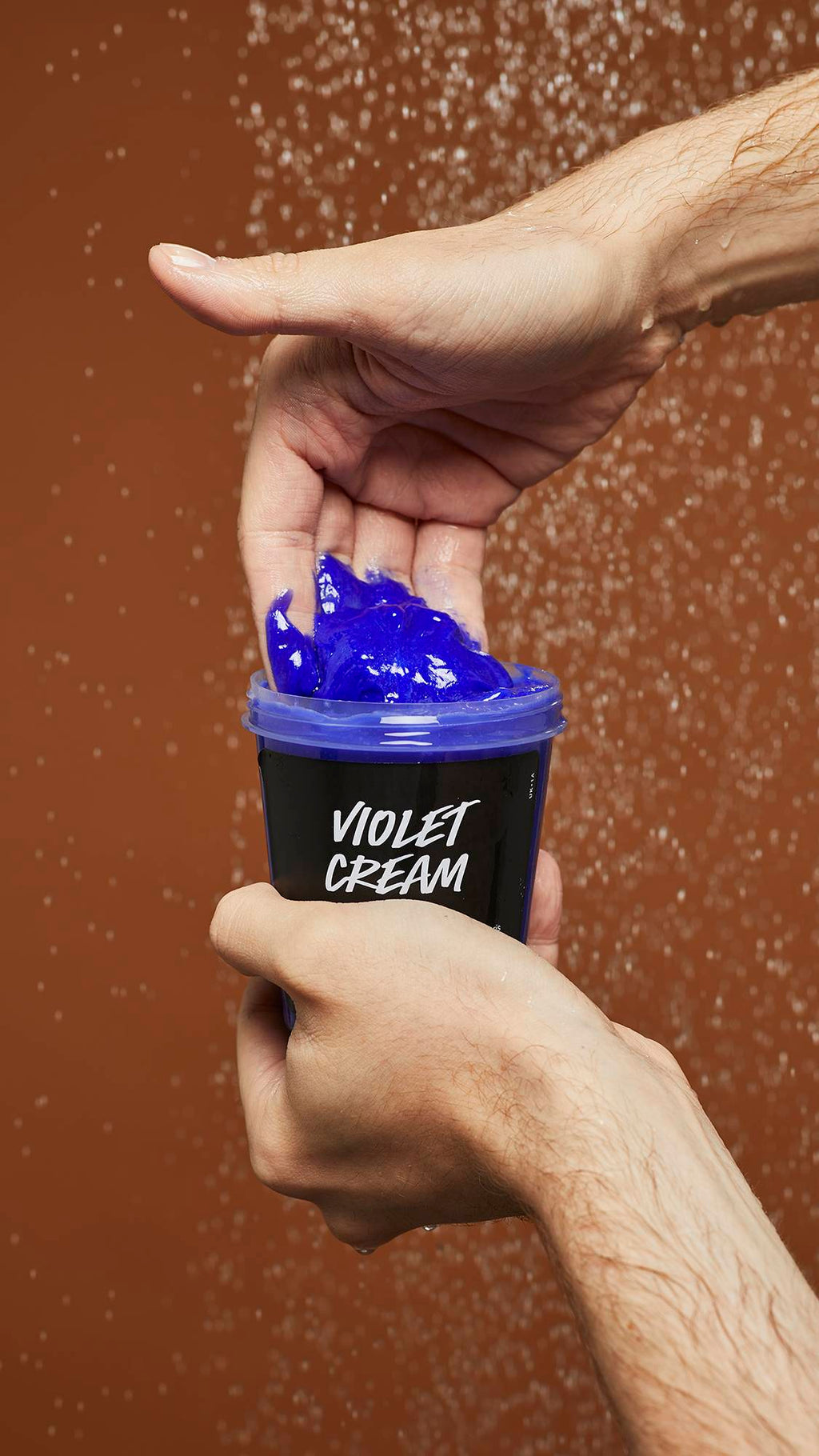 A close-up image of model scooping the vibrant Violet Cream shampoo out of the LUSH pot under running shower water.
