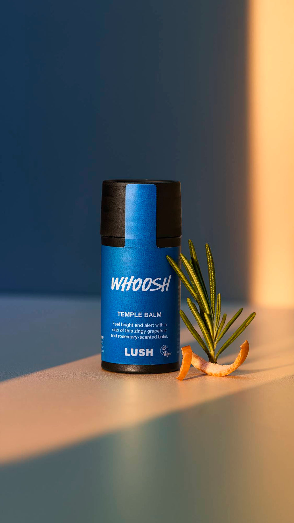 A close-up of the Whoosh temple balm surrounded by bright, natural lighting. There is a single sprig of rosemary and a twisted slice of grapefruit skin beside the product.